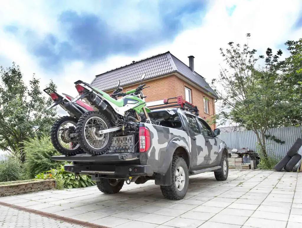 Two dirt bike motorcycles on the back of the camo truck on the driveway in residential area.