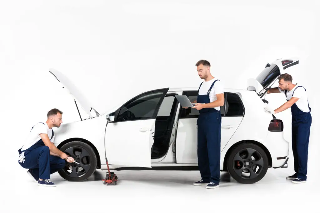 An auto mechanic changing the tire, using a laptop, and looking in an open car trunk