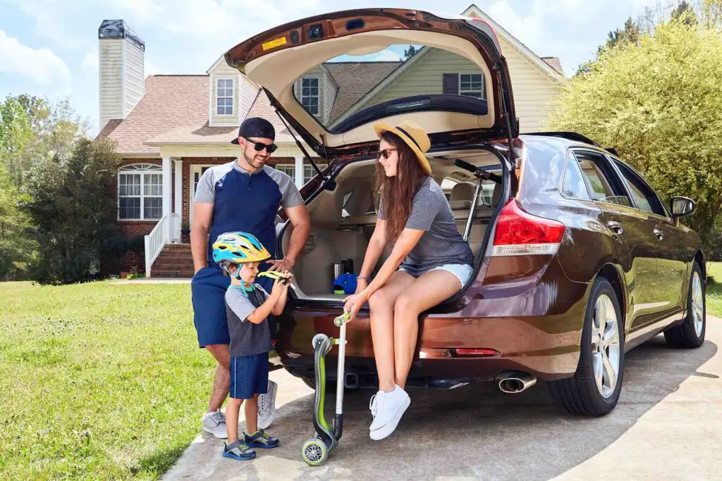 Woman sits in cars trunk and smiling,holding kickboard.Men stands near vehicle.Toddler boy wearing sun glasses.Family time.Mother,father,son near house in suburban neighborhood.Warm weather,summer.