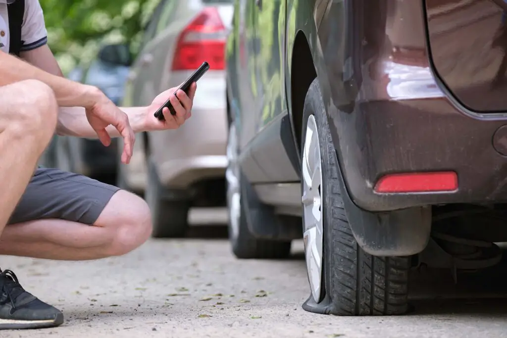 Driver calling road service for assistance having vehicle trouble with punctured flat tire on car parked on roadside.